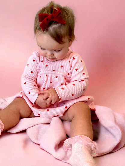 Baby girl wearing a heart-patterned dress and red bow, sitting on a pink blanket, focused on her hands.
