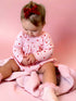 Baby girl wearing a heart-patterned dress and red bow, sitting on a pink blanket, focused on her hands.
