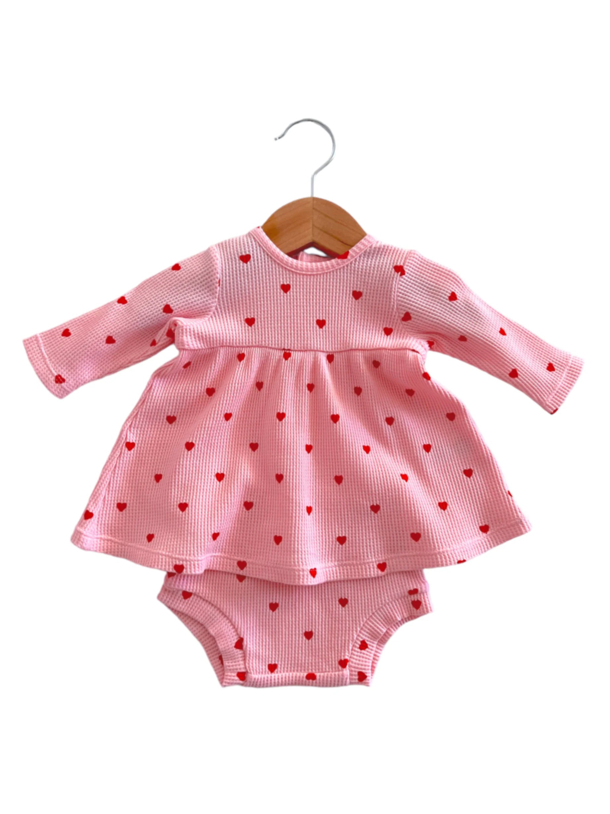 Pink dress with red heart patterns, paired with matching bloomers, hanging on a wooden coat hanger.