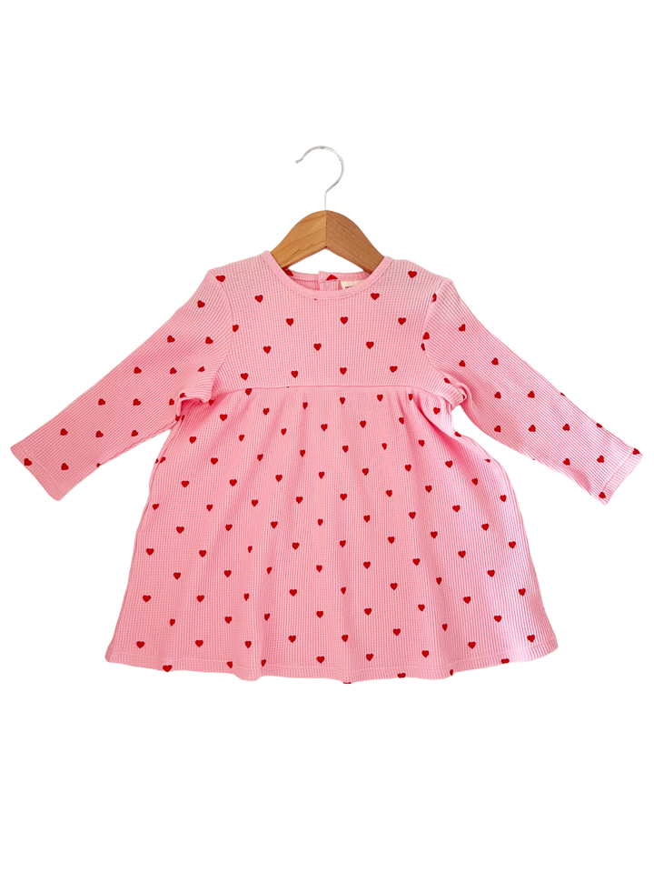 Pink dress with long sleeves and red heart patterns, hanging on a wooden hanger.