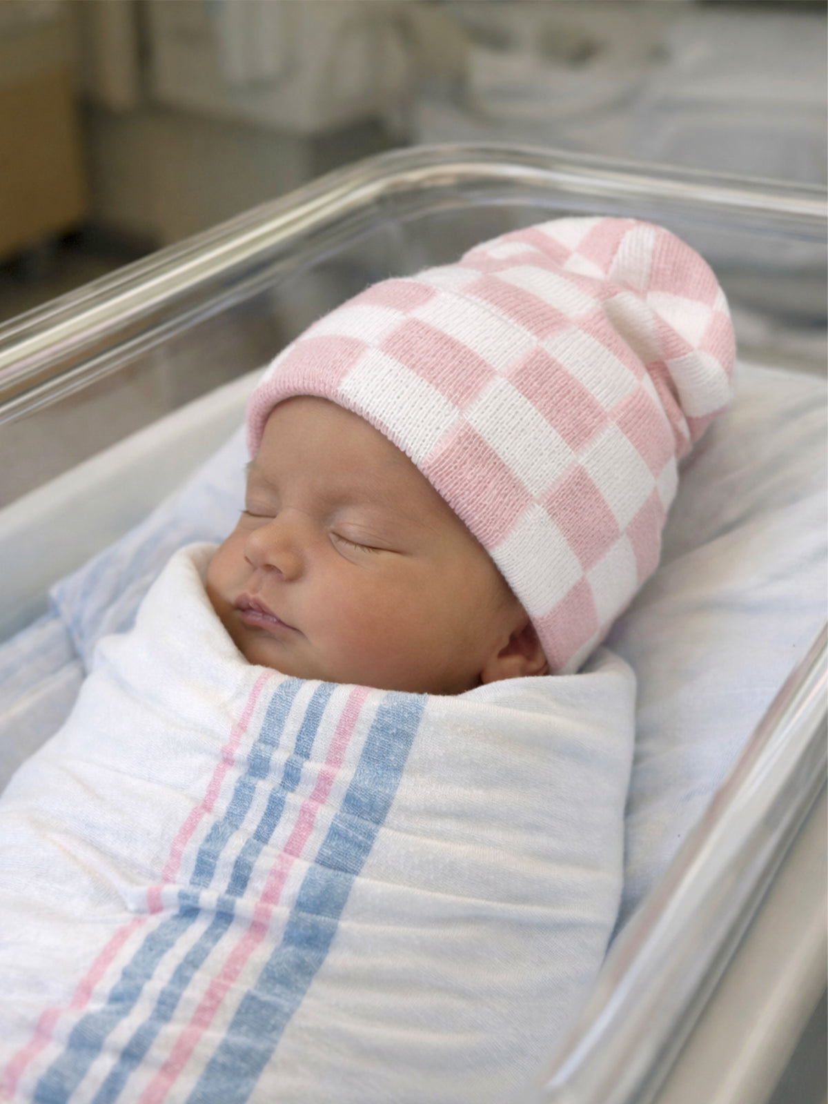 Newborn baby sleeping peacefully in a hospital bassinet, wrapped in a soft blanket with a pink and white checkered hat.
