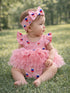 Baby girl in a pink ice cream-patterned dress and headband, sitting on grass, looking to the side.