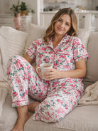 Woman in floral pajamas sits on a sofa holding a mug, smiling in a cozy, well-decorated living room.