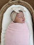 Sleeping baby wrapped in a pink checked blanket, wearing a beige bow headband, resting in a cozy basket.