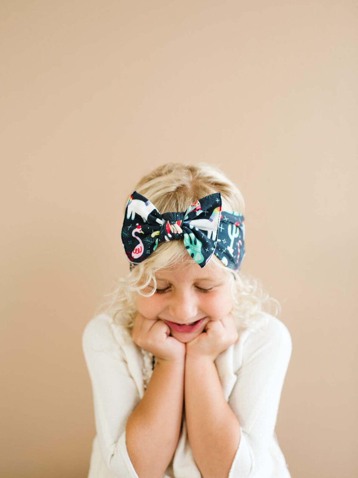 Young girl with curly blonde hair, smiling with a colorful bow headband against a soft beige background.