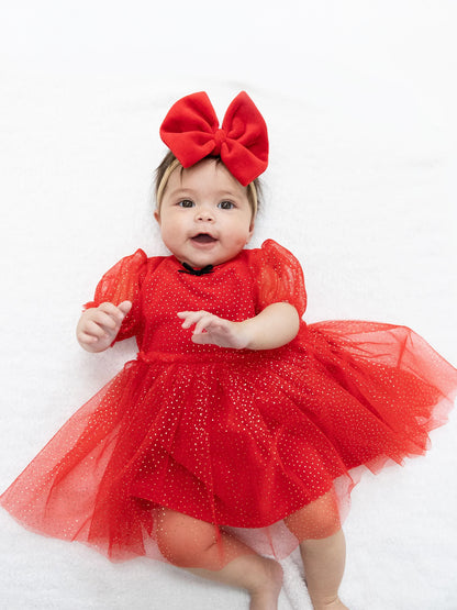 Baby in a red dress with sparkles and a large bow, smiling on a white blanket.