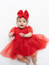 Baby in a red dress with sparkles and a large bow, smiling on a white blanket.