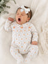 Newborn baby yawning in a white onesie with pumpkins, wearing a light bow, surrounded by soft fabrics and greenery.