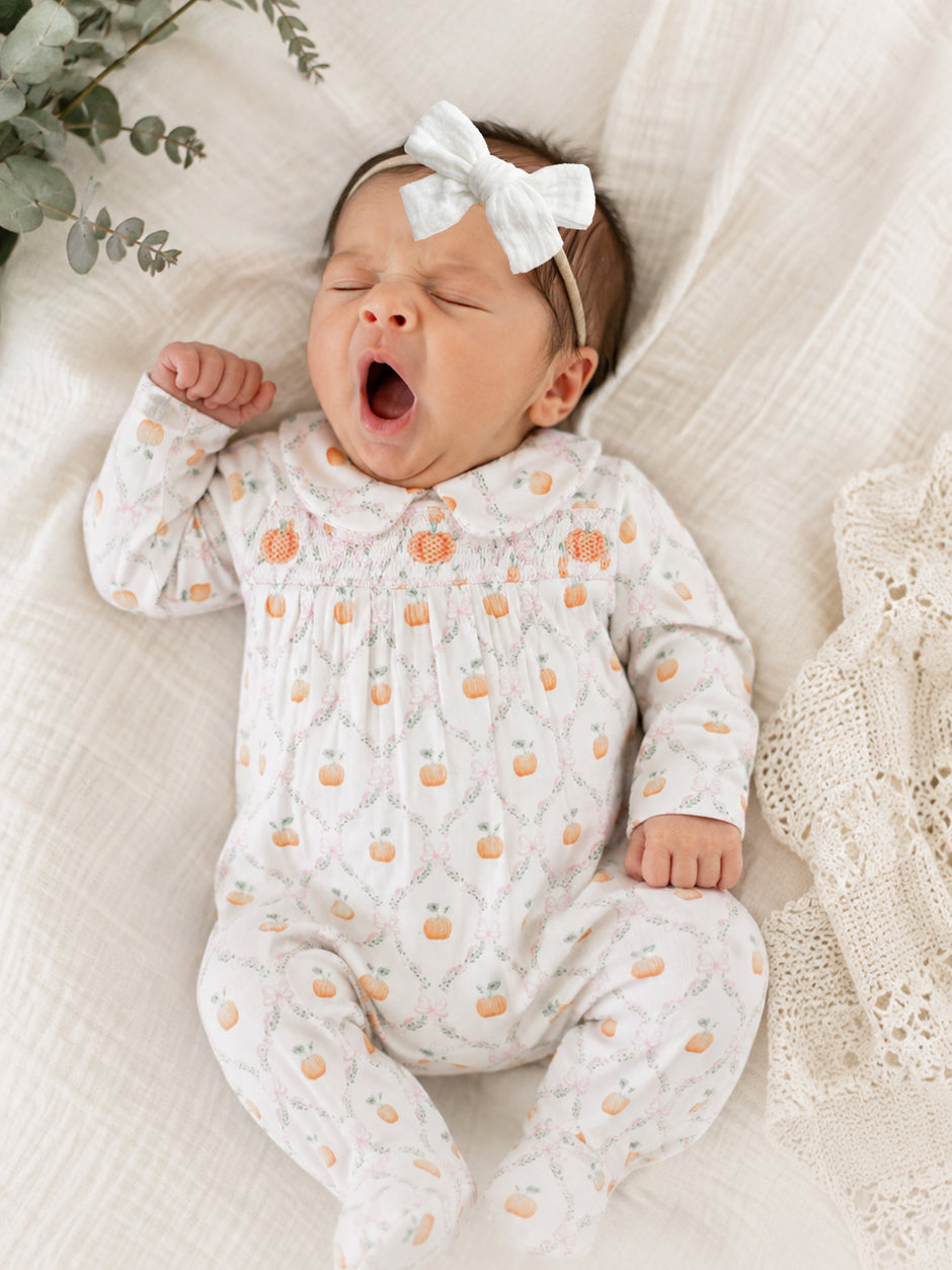Newborn baby yawning in a white onesie with pumpkins, wearing a light bow, surrounded by soft fabrics and greenery.