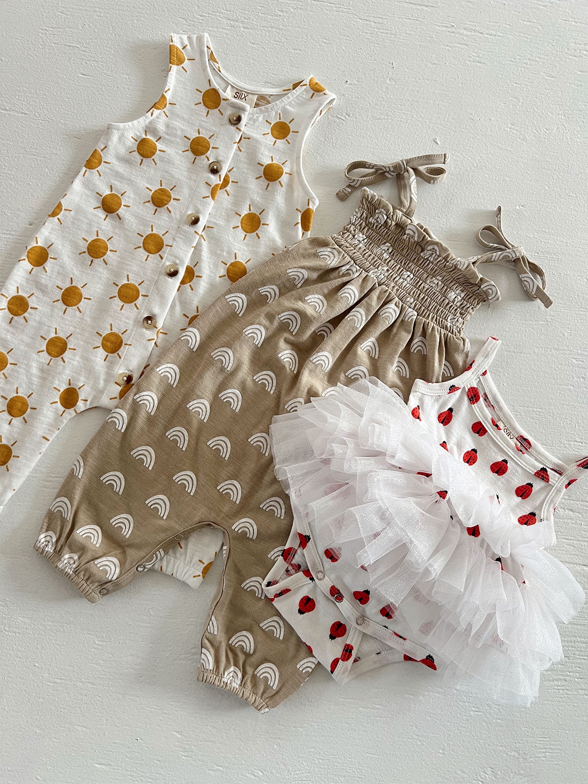 Three children’s outfits: sun-patterned dress, smocked rainbow romper, and red ladybug tulle dress against a white background.