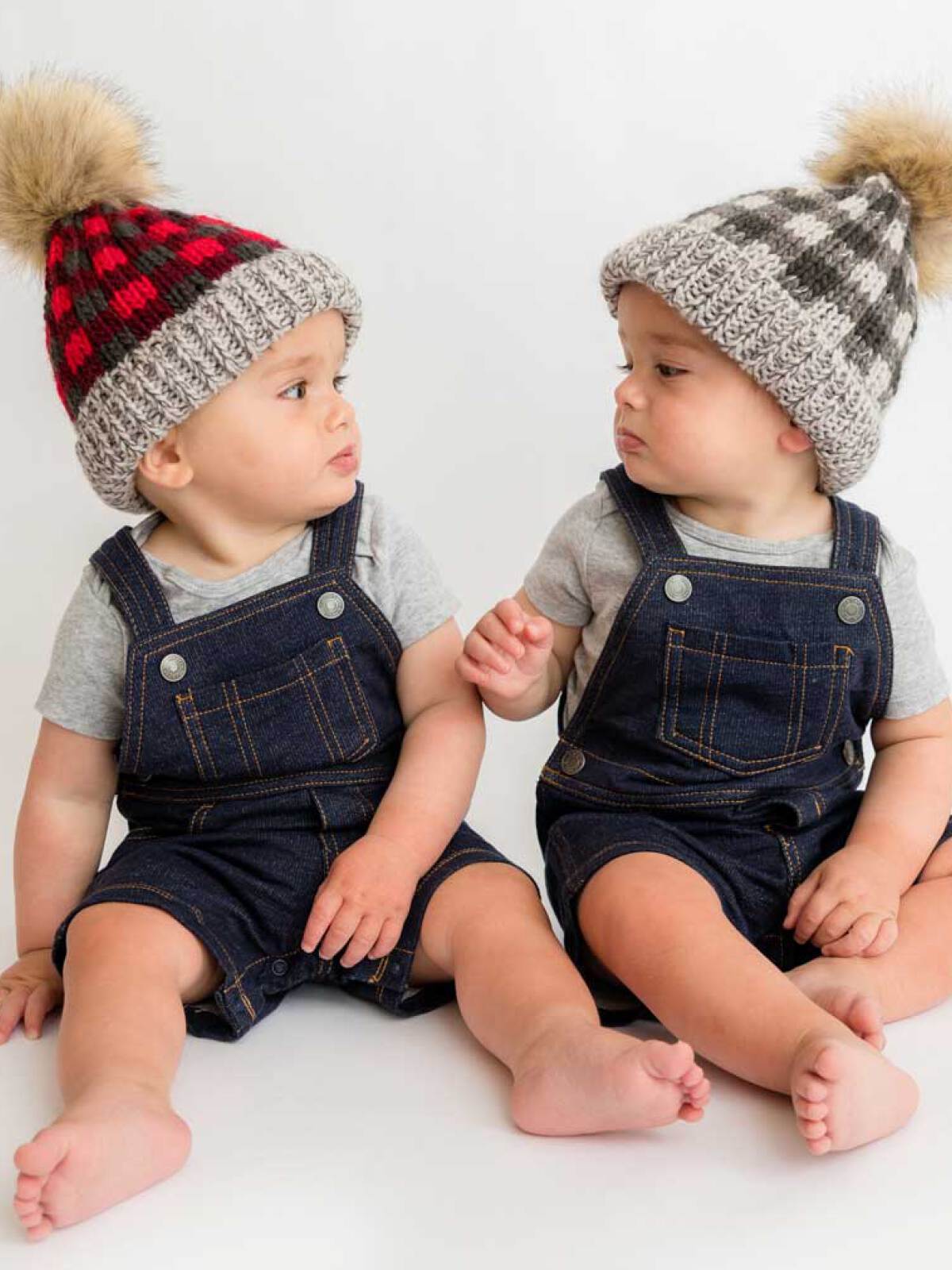 Two babies in denim overalls and knitted hats sit side by side, interacting with each other on a light background.