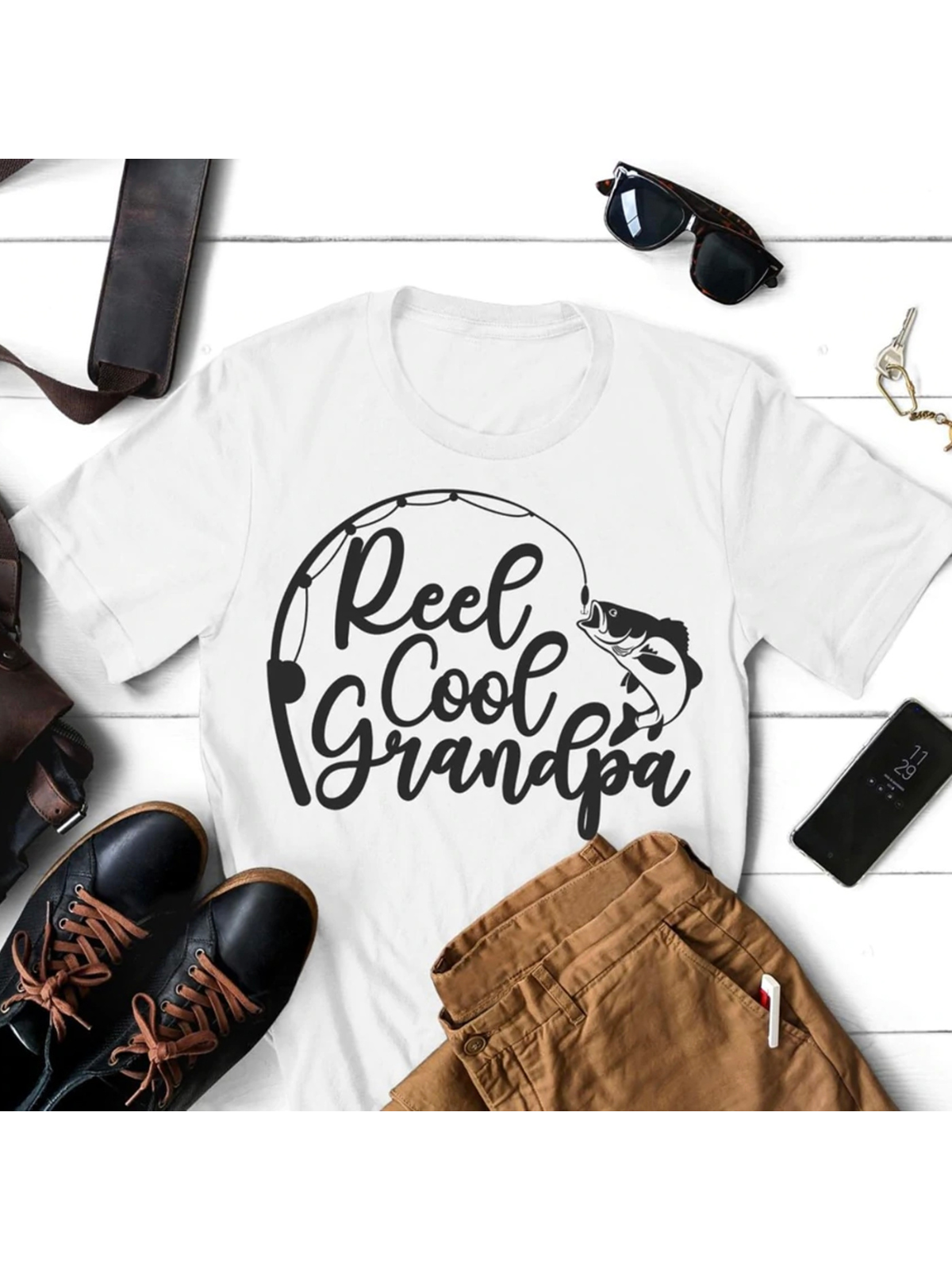 White t-shirt with "Reel Cool Grandpa" text and fishing graphic, surrounded by casual accessories and clothing.