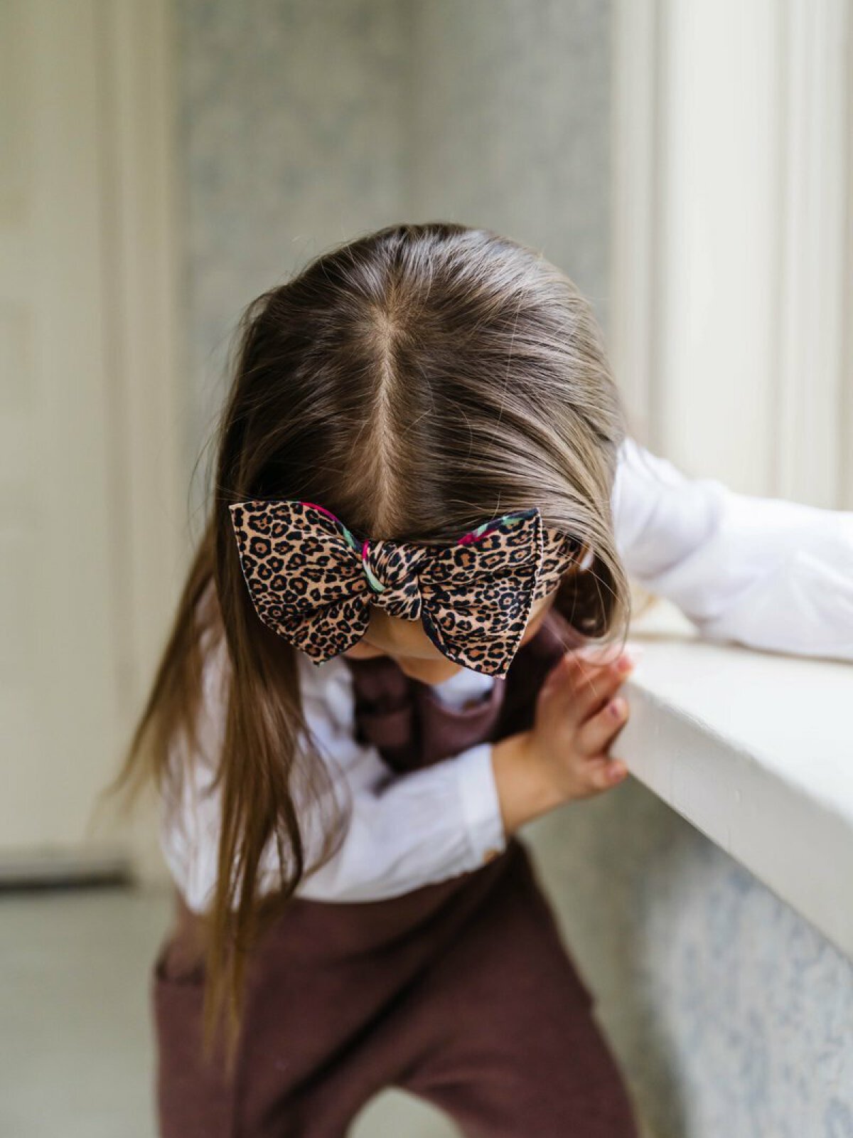 Child with long hair wearing a leopard-print bow, leaning against a windowsill in a softly lit room.