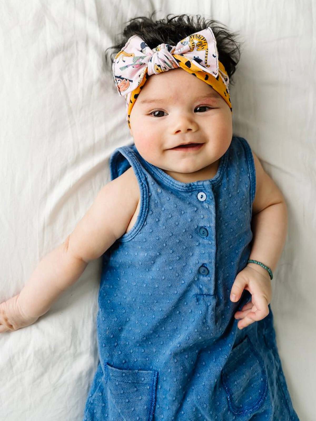 Smiling baby in a blue dotted dress with a colorful headband, lying on white bedding.