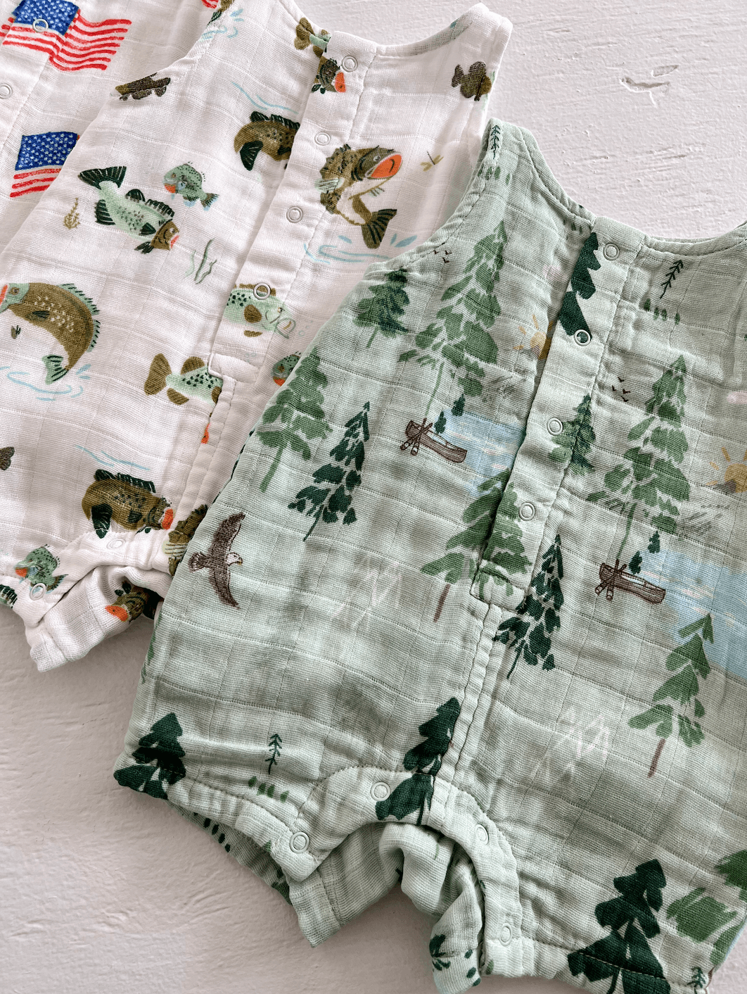 Three baby onesies with nature and fish prints in varying colors, laid out on a light background.
