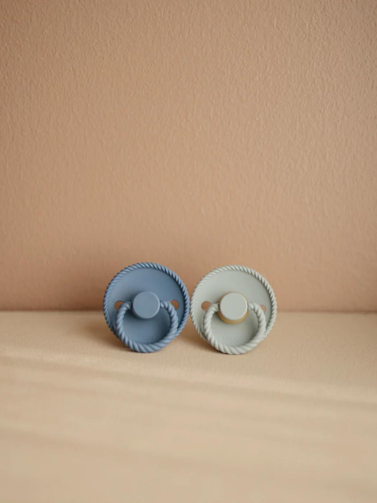 Two pacifiers in blue and green colors on a beige background, showcasing a simple, modern design.