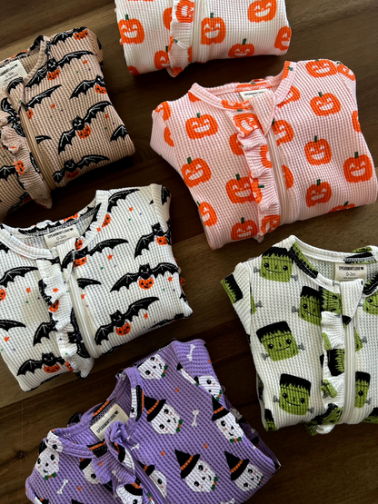 Colorful kids' Halloween-themed pajamas featuring pumpkins, bats, ghosts, and monsters laid out on a wooden surface.