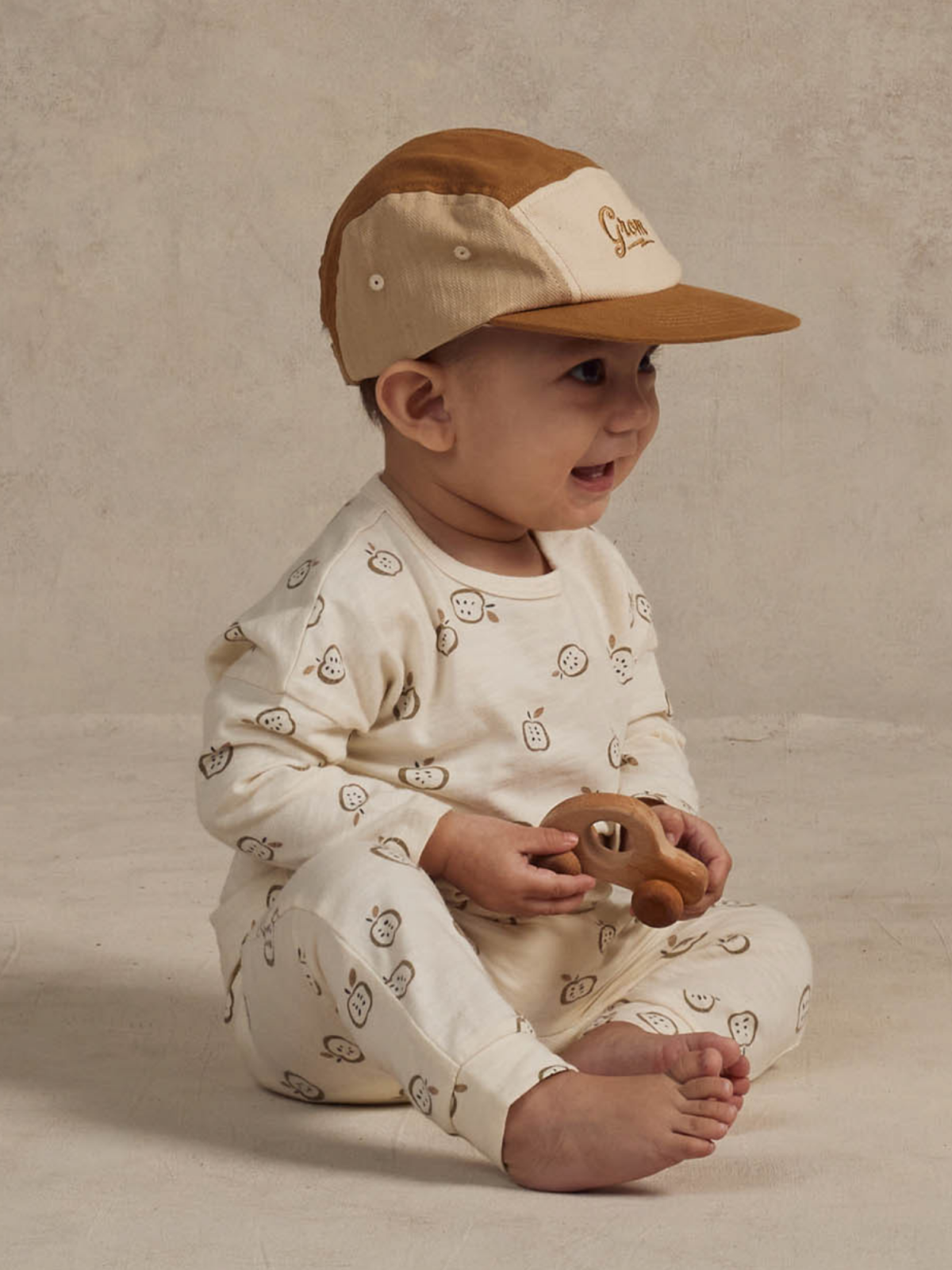 Smiling baby in a patterned pajama holding a wooden toy, wearing a beige and brown cap, sitting on a neutral background.