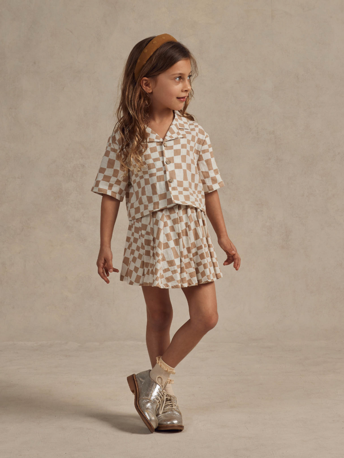 Child in a checked outfit with a matching headband, silver shoes, against a neutral background.