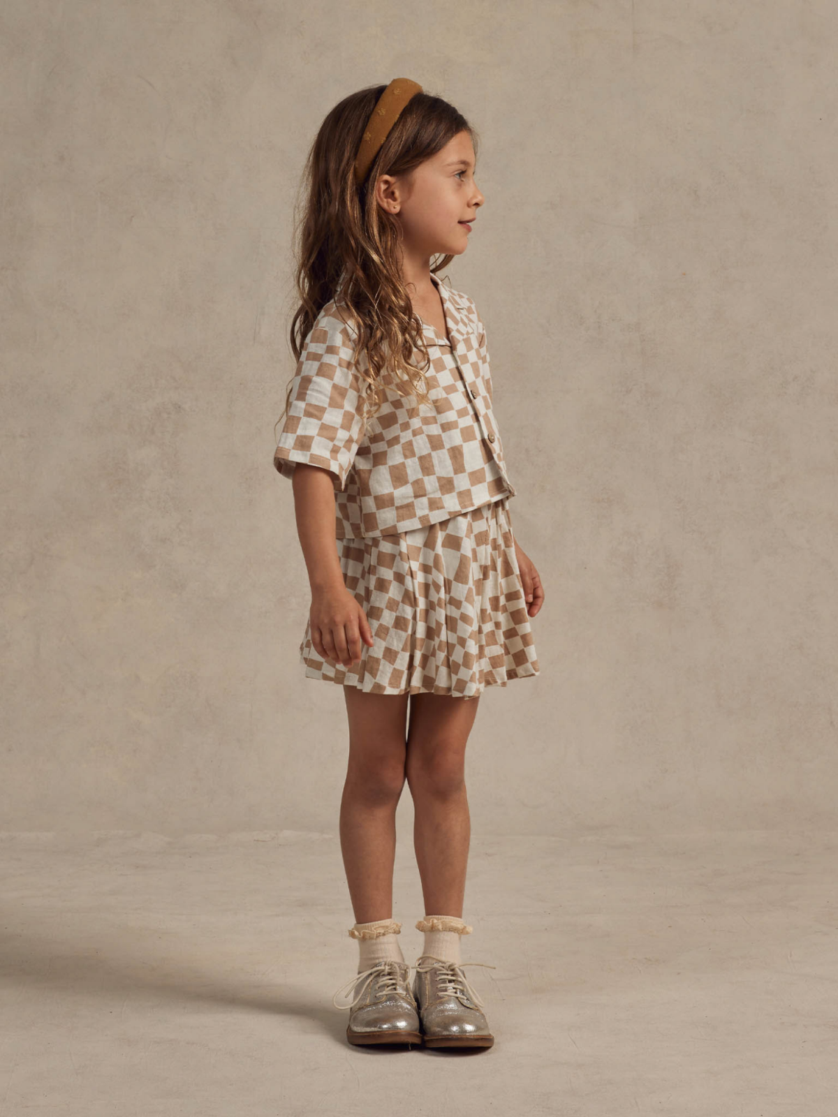Girl with long hair in a checkered outfit, standing against a neutral backdrop, wearing shiny shoes.
