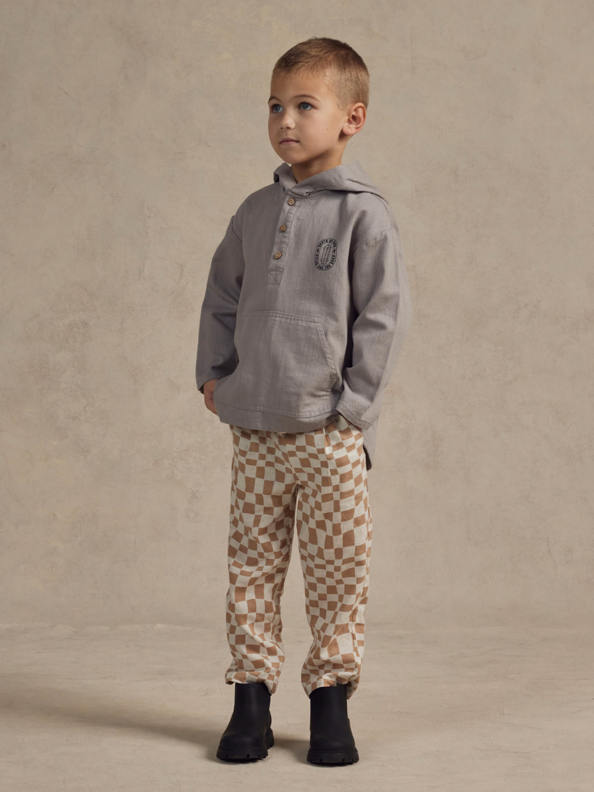 Young boy wearing a gray hooded shirt and patterned beige pants, standing on a neutral background.