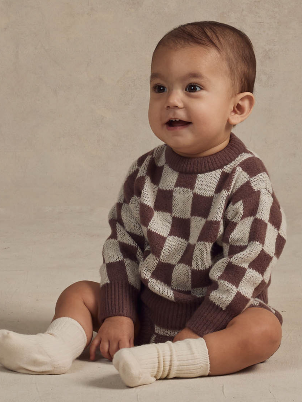 Smiling baby in a checkered sweater sits on a neutral background, wearing white socks.