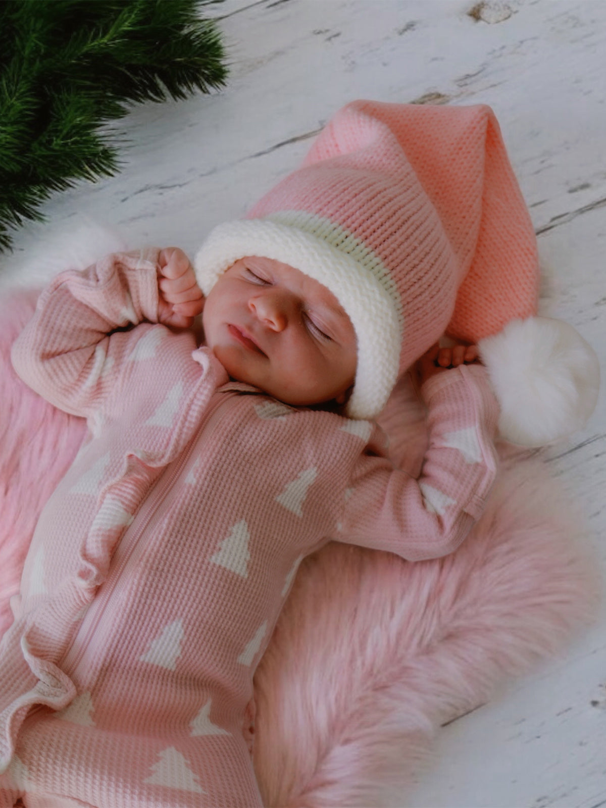 Newborn in a pink outfit and festive hat, sleeping on a soft pink blanket with greenery in the background.