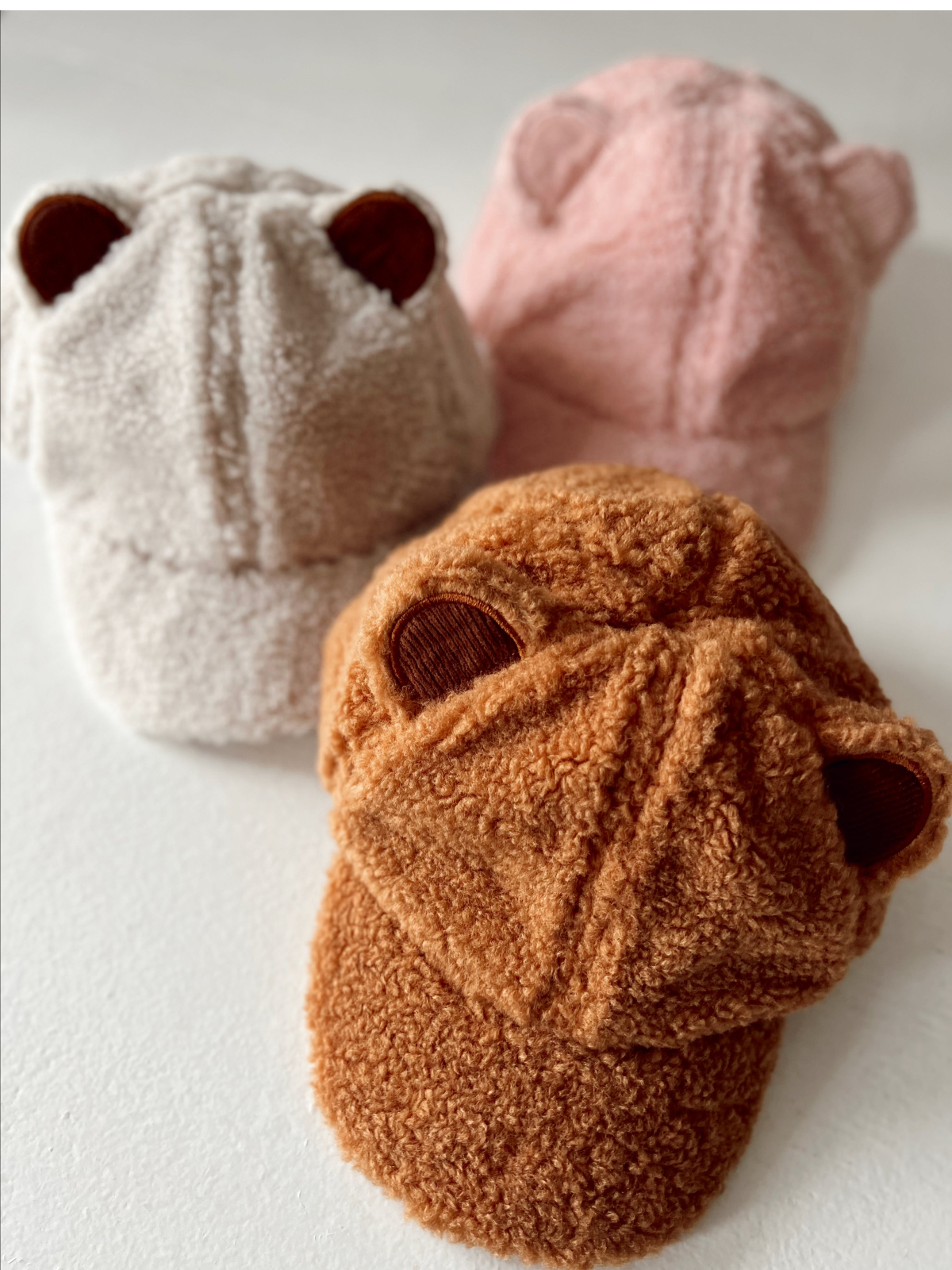 Three cozy hats with ear details in white, pink, and orange, displayed on a white surface.