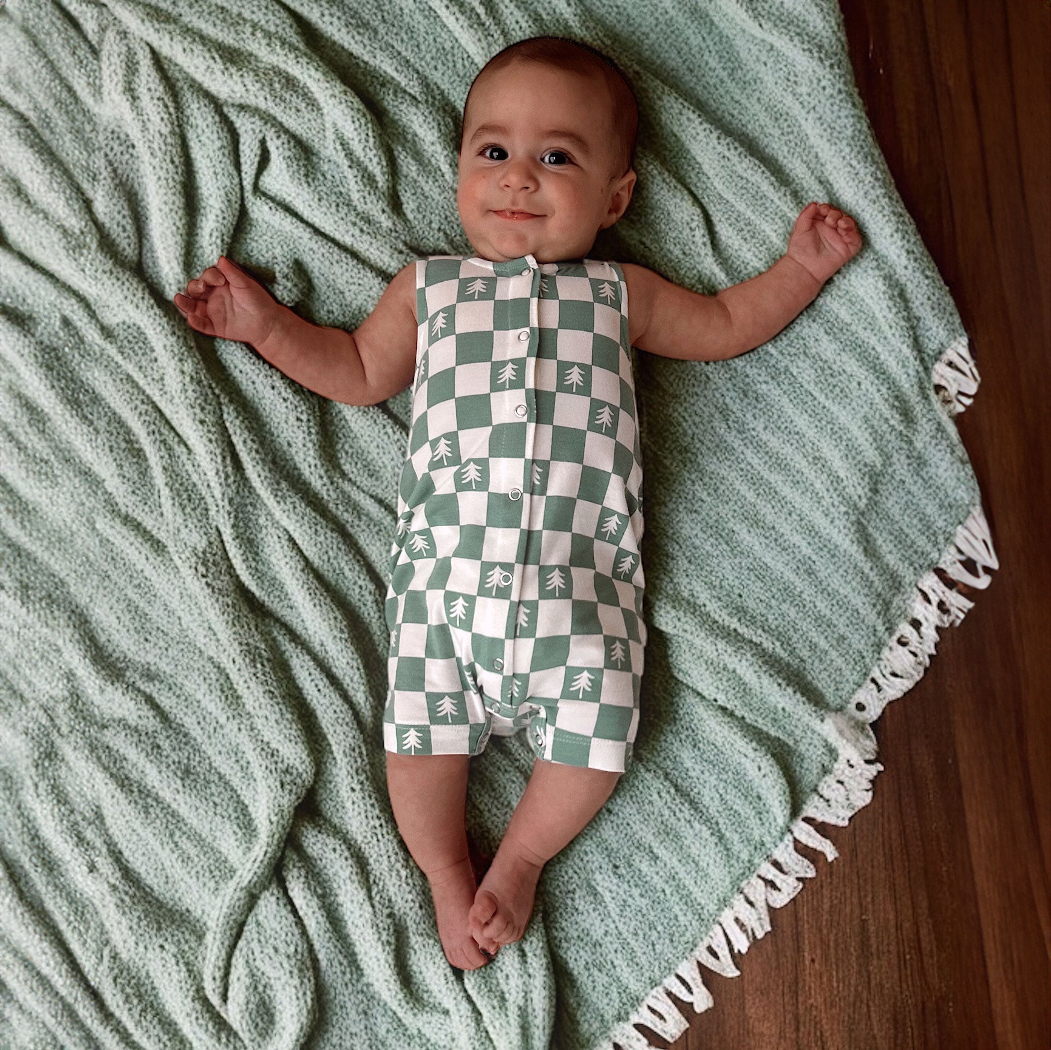 Infant lying on a green blanket, wearing a checkered onesie with tree patterns, smiling at the camera.
