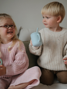 Two smiling children in cozy sweaters, enjoying drinks together on a sofa.