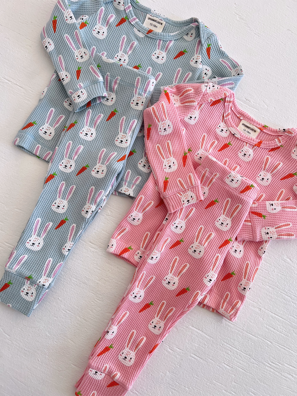 Two sets of children's pajamas featuring bunny and carrot patterns, one in blue and one in pink.