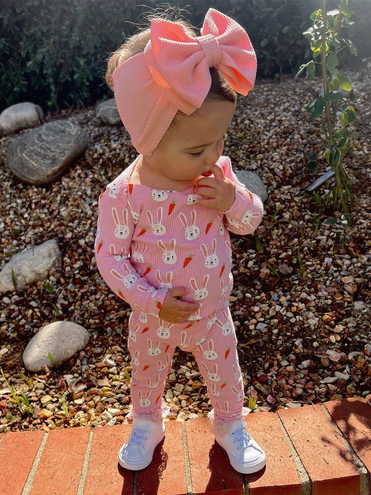 Toddler wearing bunny-patterned outfit and large bow headband, standing on rocky ground with greenery in background.