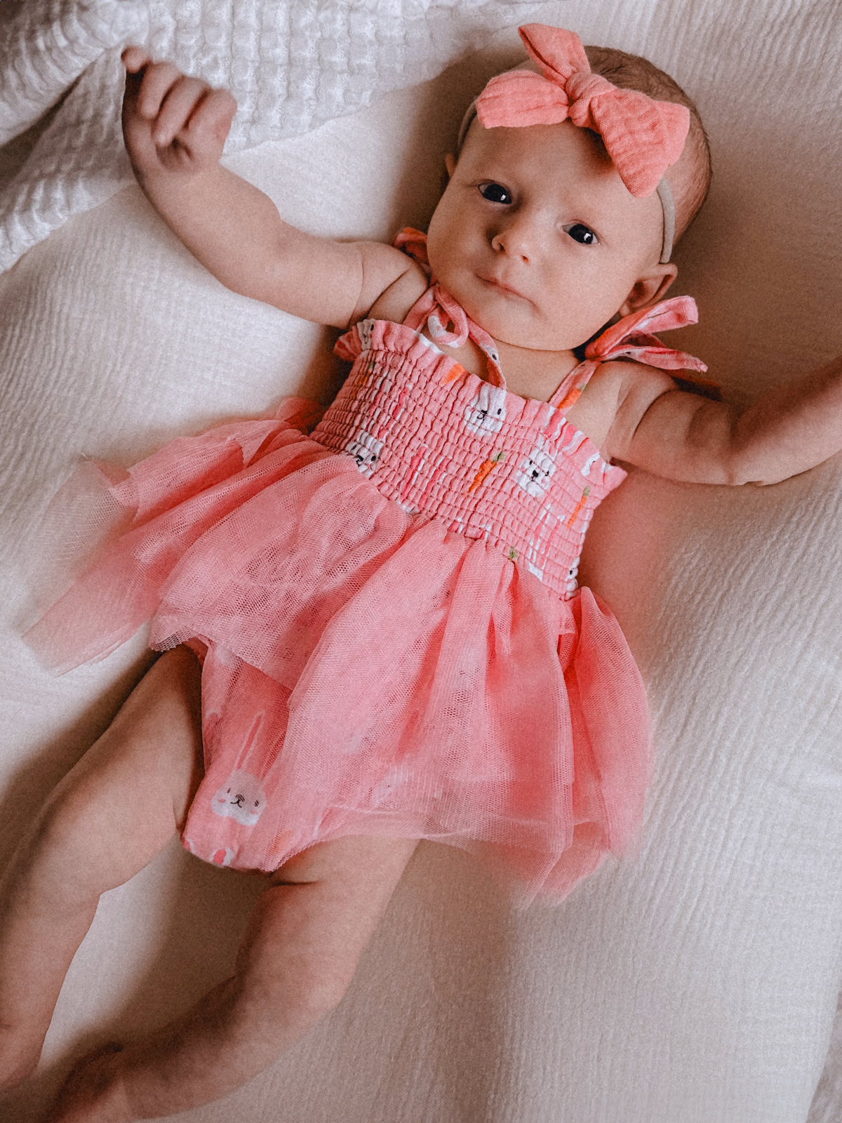 Baby in pink dress with bunny print, wearing a matching headband, lying on a textured white blanket.