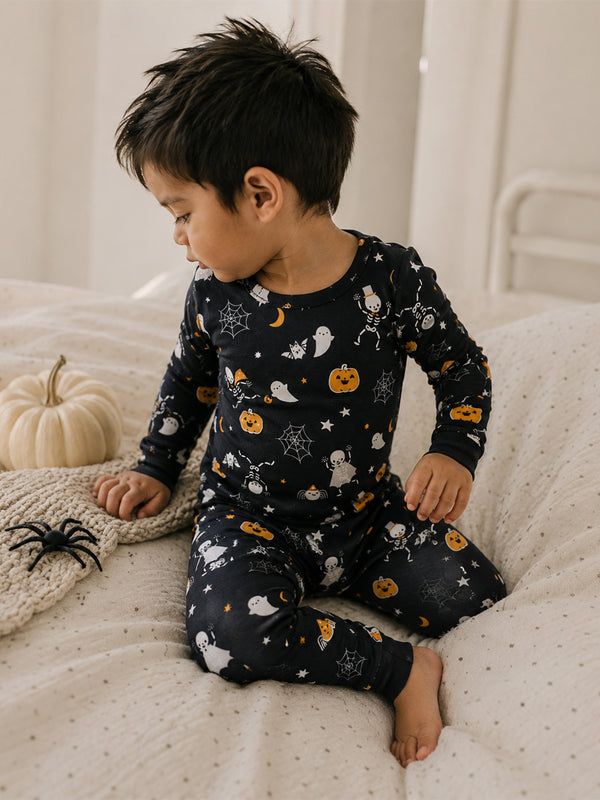 Child in Halloween-themed pajamas with ghosts and pumpkins, sitting on a bed next to a decorative pumpkin and spider.