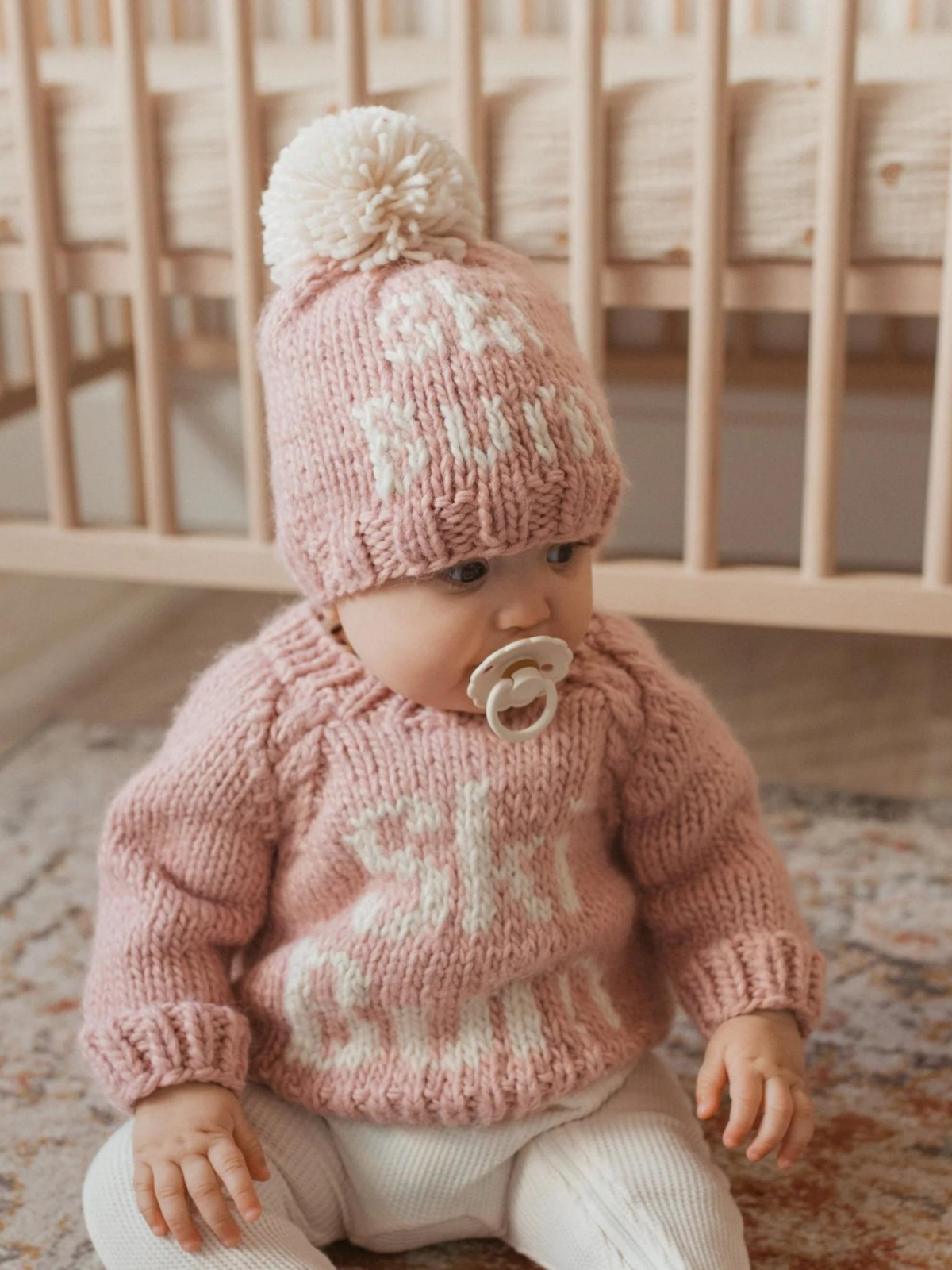 Baby in a knitted pink sweater and matching hat, sitting on a patterned rug near a crib, with a pacifier in mouth.