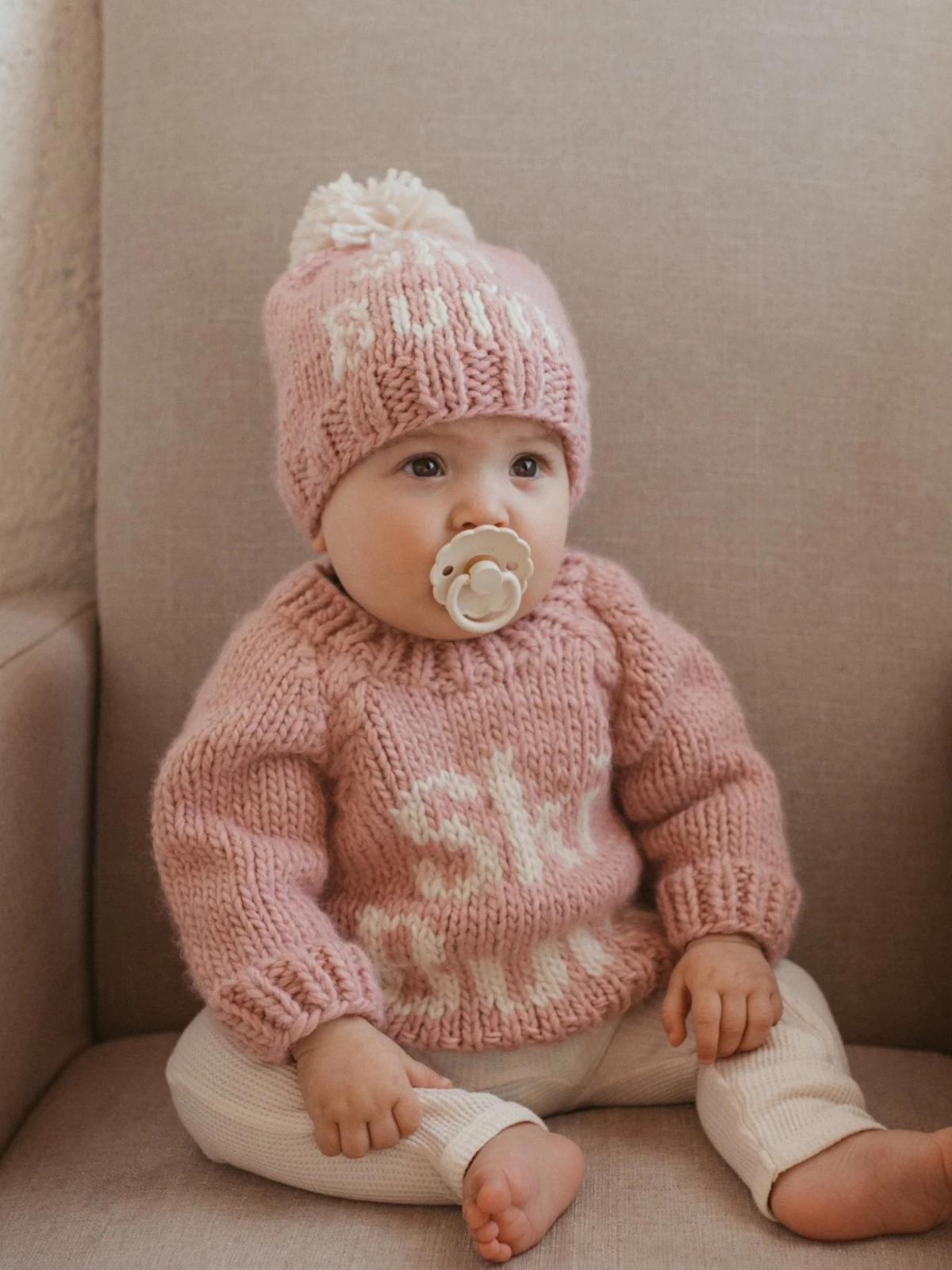 Baby in a pink knitted sweater and hat with pom-pom, sitting on a beige chair, holding a pacifier.