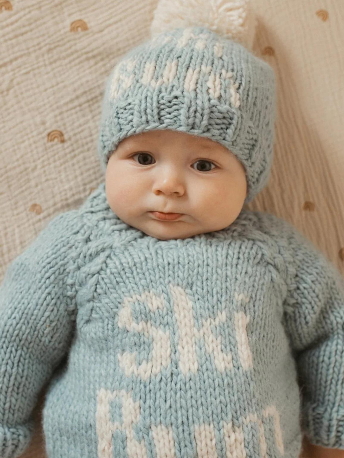 Baby in a knitted blue "Ski Bum" sweater and matching hat, sitting on a light textured blanket.