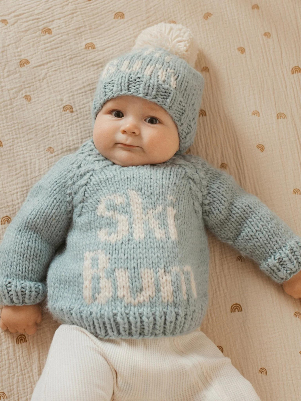 A cute baby wearing a light blue "Ski Bum" sweater and matching hat, resting on a patterned blanket.