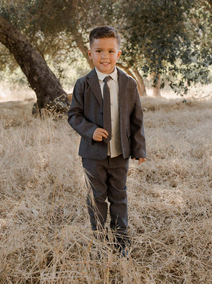 Smiling young boy in a suit standing in a grassy field, surrounded by trees.