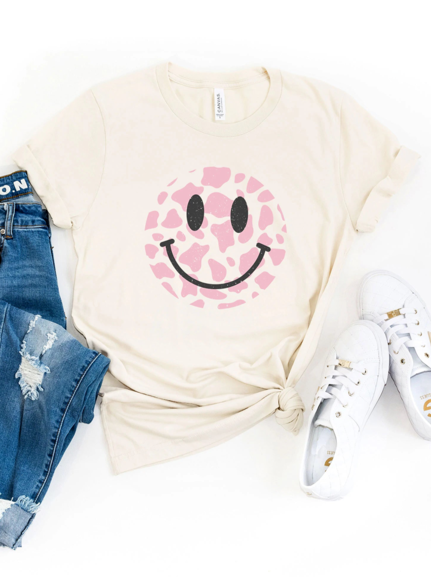 Cream T-shirt with a pink giraffe-patterned smiley face, styled with blue jeans and white sneakers.