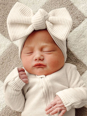 Newborn baby wearing a white outfit and large bow, peacefully sleeping on a soft, patterned blanket.