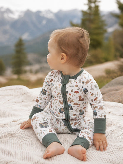 Toddler in nature-themed pajamas sitting on a blanket with mountains in the background.