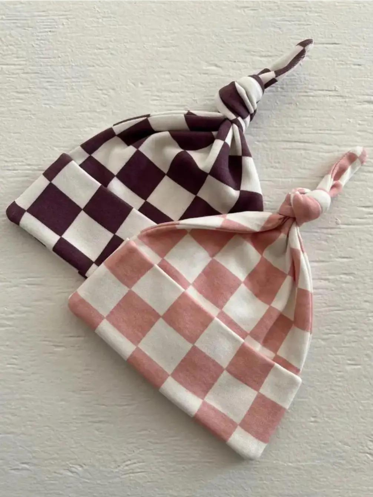 Two checkered baby hats in maroon and pink, featuring knotted tops, resting on a textured white surface.