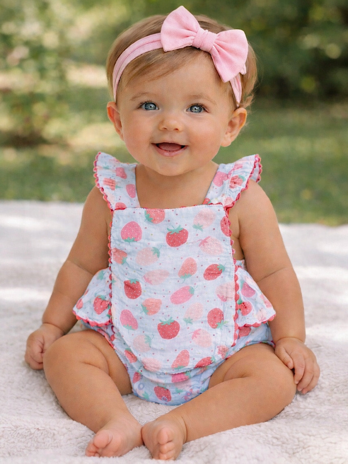 Smiling baby in a strawberry-patterned outfit and pink headband, sitting on a soft rug outdoors.