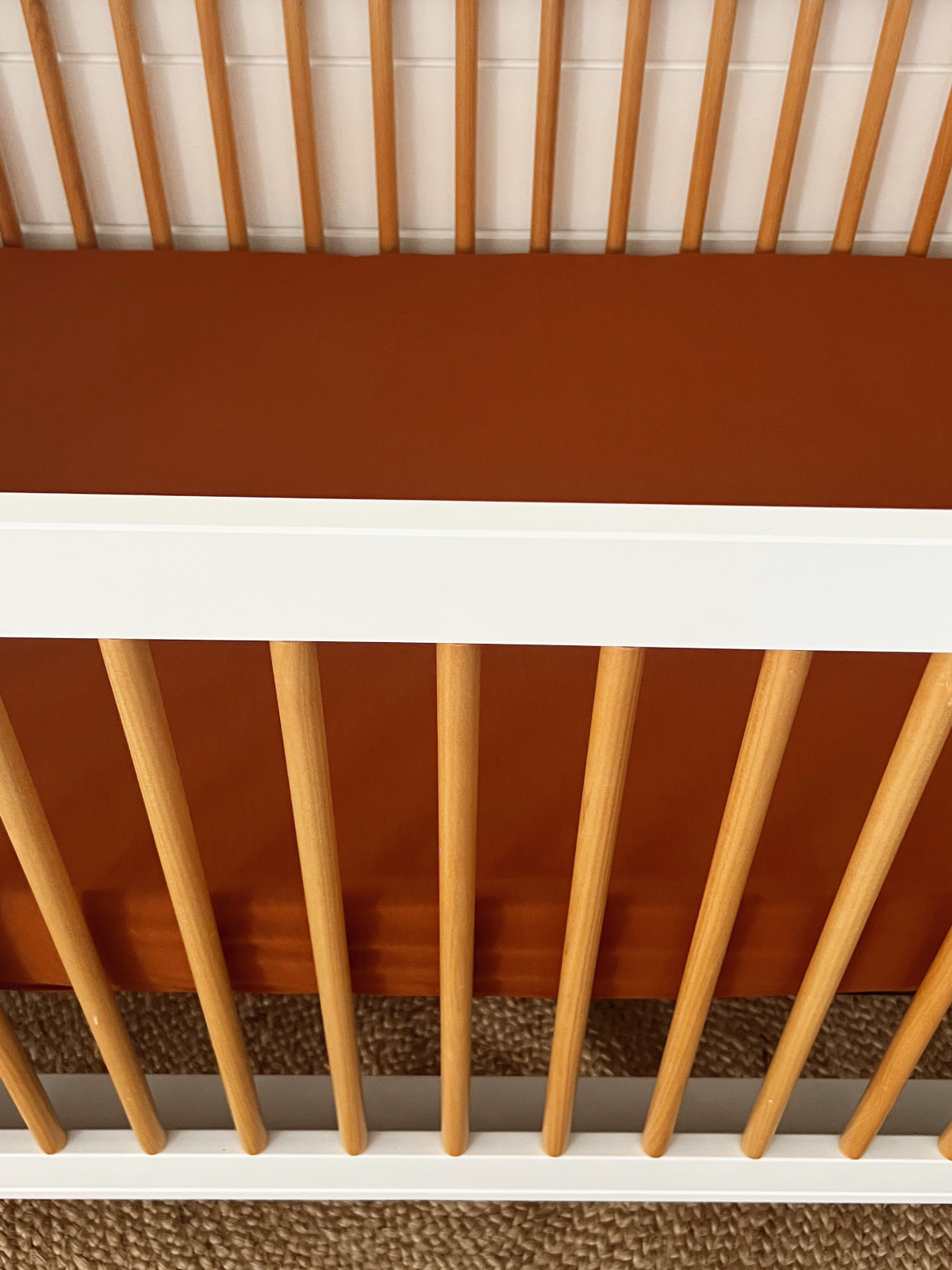 Baby crib with wooden slats and orange bedding on a woven rug.