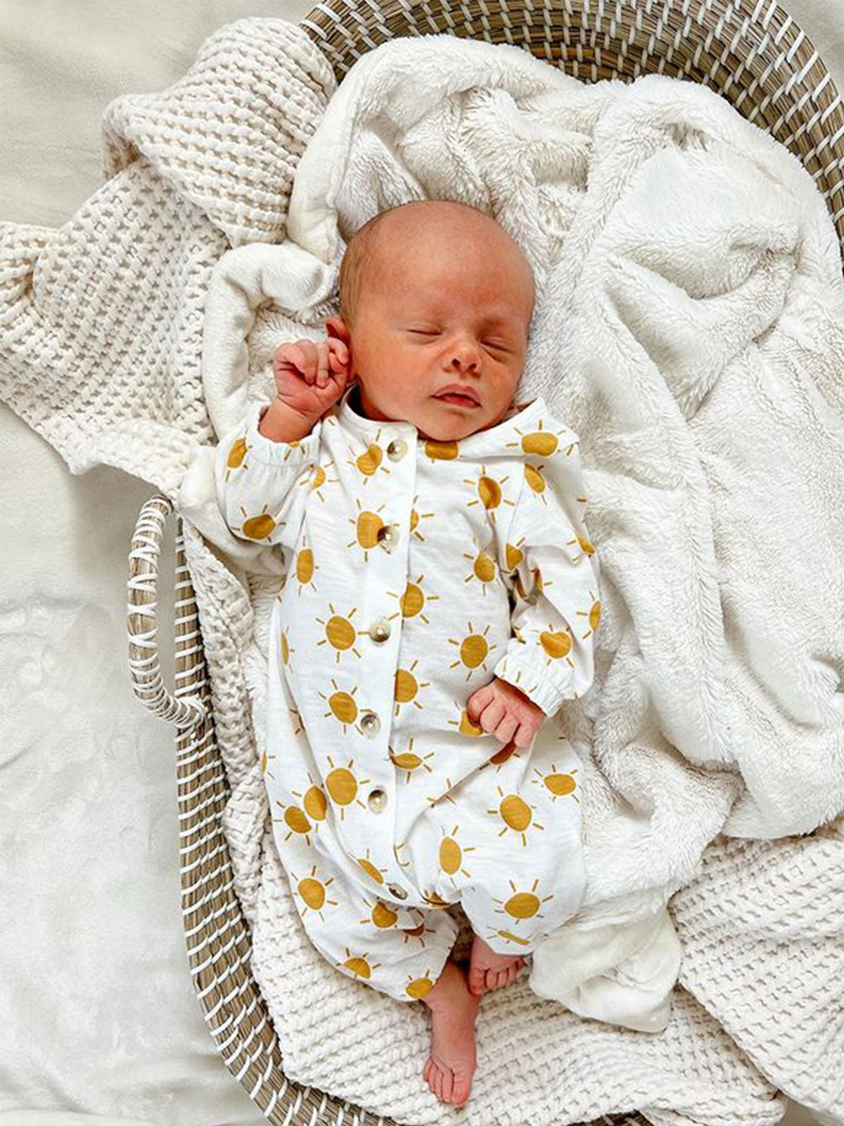 Baby in a sun-patterned onesie peacefully sleeping in a cozy woven basket surrounded by soft blankets.