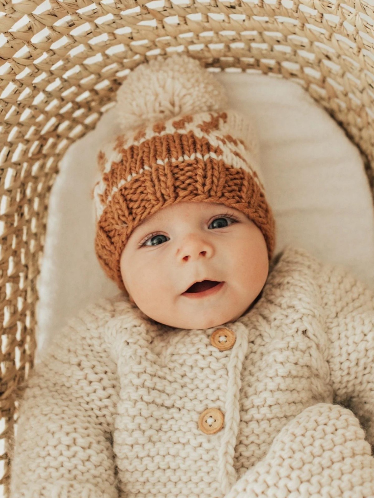 Smiling baby in cozy knitted sweater and hat, nestled in a woven basket.