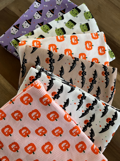 Colorful Halloween-themed kitchen towels featuring pumpkins, ghosts, and bats stacked on a wooden surface.
