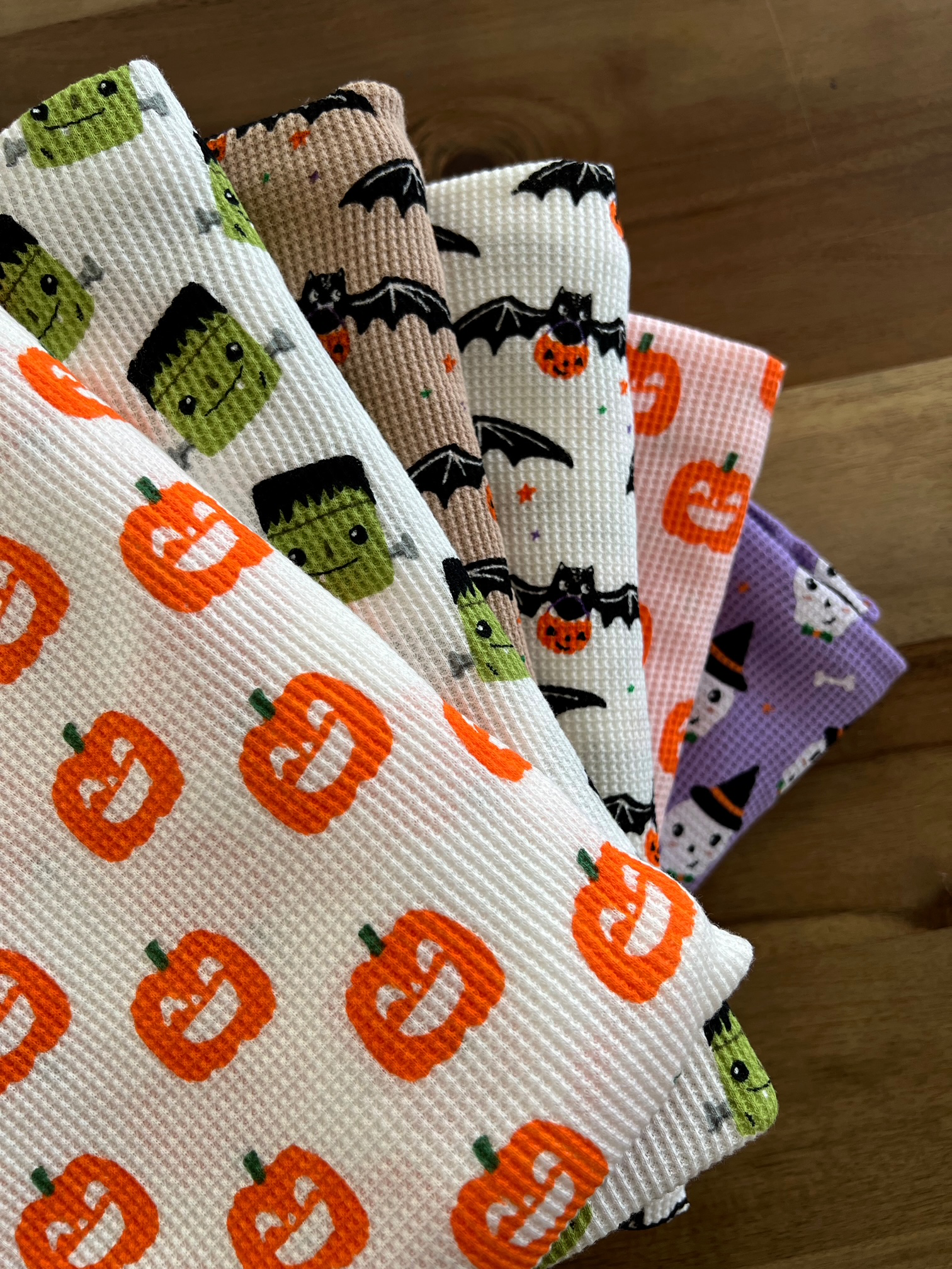 Colorful Halloween-themed dish towels featuring pumpkins, bats, and spooky characters on a wooden surface.