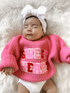 A serene baby girl in a pink "Sweet Heart" sweater and a white headband, resting on a fluffy white blanket.